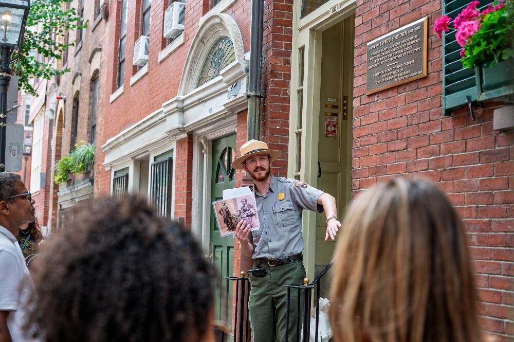 National Park Service ranger in front of brick townhouse holding black and white image of 19th century abolitionist Bostonians.