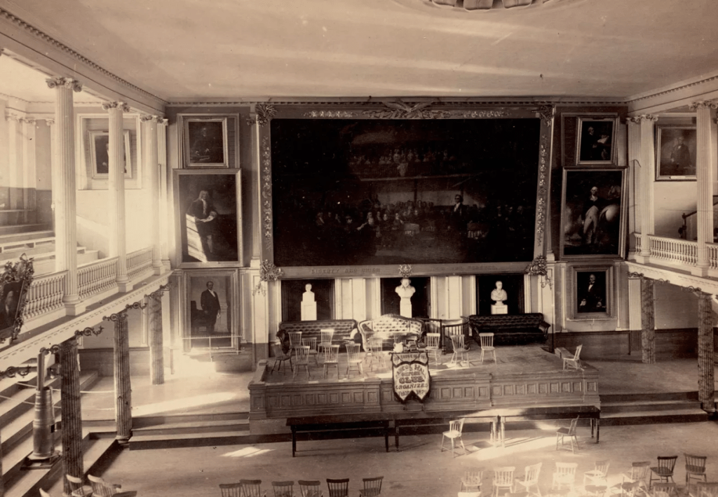 Black-and-white interior view of Faneuil Hall depicting large-scale paintings and portraits on back wall, decorative columns to the left, and a stage facing sets of chairs in the foreground.