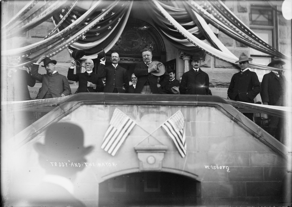 A group of men in formal attire stand on a decorated balcony with US flags, some tipping their hats to a crowd below.