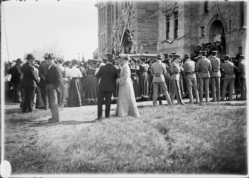 A crowd of people, including men in uniforms and women in dresses, gathers outside a decorated stone building for an event.