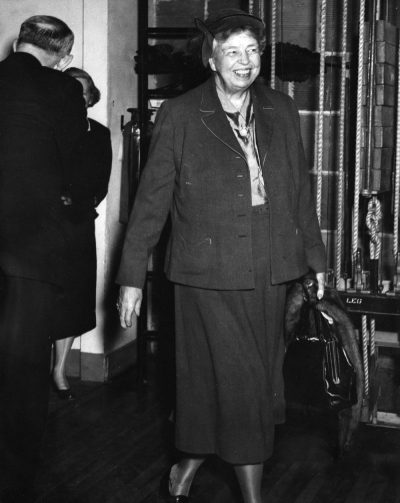 An older woman in a dark suit walking indoors with a smile, holding a handbag.