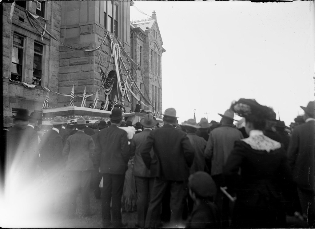 Black and white image of people gathered facing a decorated stone building, featuring American flags and a speaker on a platform.